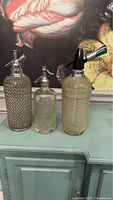 Photo showing three vintage seltzer bottles with chain link metal coverings and glass bodies placed on a greenish wooden surface with a painting background.