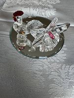Clear Swarovski crystal rooster figurine with red comb and pink-accented flower figurine on round mirror base placed on patterned tablecloth.