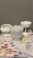 Three white milk glass vases from front view showing shapes, scalloped tops, and fluted bottoms on a patterned cloth background.
