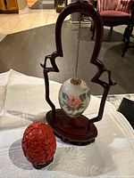 White egg with hand painted floral and bird designs hanging on a carved rosewood stand, red carved cinnabar egg on black stand beside it.