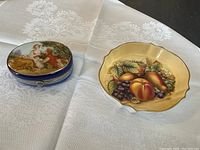 Both vintage trinket holders shown side by side on white embroidered cloth: oval blue trinket box with romantic Rococo scene and round scalloped fruit bowl