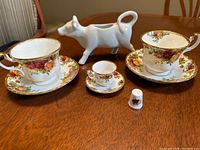 Photo showing two floral patterned Royal Albert teacups with matching saucers, a mini cup and saucer, white cow milk pitcher, and a small thimble all displayed on wooden table.