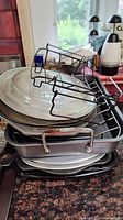 Stacked baking pans including square, rectangular, and round shapes with a glass lid on top and a metal wire cooling rack beside and on top