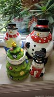 Four ceramic snowman-themed figures on windowsill: three cookie jars and one piggy bank. Various sizes and colors including a green Christmas tree cookie jar. Visible chips on one snowman jar's nose and tree ornament star.