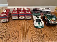 Seven Hess vehicles lined up on the floor including four red fire trucks, one white and green race car, and one green rescue truck.