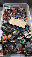 Top view of a plastic container filled with assorted LEGO blocks, plates, and pieces in various colors and sizes including tan baseplates and multi-colored bricks.