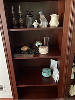 Wooden display cabinet with two miniature oil lamps, figurines, coasters, clocks, and vases on multiple shelves.