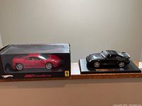 Photo showing a red Ferrari 360 Modena model car inside its original black box and a black Porsche 911 Turbo model car on a display base.