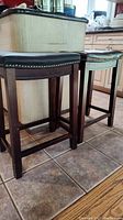 Side view showing two different countertop height stools, each with dark wooden legs and black upholstered seats. One stool has nail head detailing along seat edge.