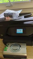 Black HP OfficeJet Pro 6968 printer with touchscreen, extra cartridges in plastic bag on top, and silver Canon CP220 compact photo printer below.