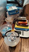 Weight Watchers digital scale, PlayStation 2 memory card, cassette tapes, CD-R discs, vaporizer, glass paperweight, and tin basket arranged on a wood surface