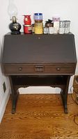 Full view of the vintage wooden secretary desk with the slant front closed. Several candles and a decorative lamp placed on top.