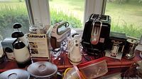 Overview of small kitchen appliances on a tiled surface near window, including Cuisinart Mini-Prep food processor box, Oster hand mixer, Black & Decker toaster, two espresso percolators, waffle maker, electric cheese grater, KitchenAid hand mixer, and cutlery sharpener.