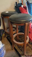 Photo showing two wooden bar height stools with round, padded swivel seats featuring brass nailhead trim, missing some trim pieces. Stools have four legs connected by circular footrests and show wear.