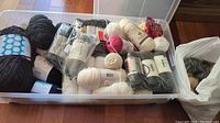 Wide view inside transparent plastic bin showing an assortment of yarn skeins in black, white, cream, gray, dark blue, pink, and patterned varieties along with a white plastic bag containing faux fur pompoms.