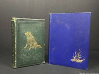 Front covers of two antique books showing 'The Wild North Land' green cover with a seated dog illustration and 'The Wooden Walls Among The Ice Floes' blue cover with a ship in gold.