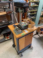 Wide angle showing drill press mounted on a cabinet with drill bit cases and basket on top, in a workshop environment.
