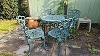 Wide view of one round cast metal table surrounded by four matching cast metal chairs on stone patio, weathered green finish.