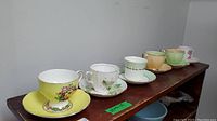 Six vintage English china teacups and matching saucers arranged on a wooden shelf. Colors are predominantly pastel with floral detailing and gold trim accents.