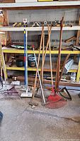 Several long-handle gardening tools arranged against yellow shelves including rakes, hoes, and digging tools with some decorative yard art and containers on shelves.