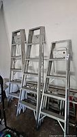Three Lite folding step ladders standing upright in a garage. One ladder is shorter (approx. 5 feet), two ladders are taller (approx. 6 feet).