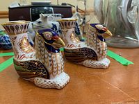 Pair of Royal Crown Derby bird-shaped candlestick holders showing detailed hand-painted designs in gold, blue, red, and white colors, placed on a wooden surface with other ceramics in the background.