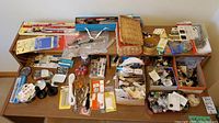 Top view of a large assortment of sewing notions on a wooden table including several containers with buttons, scissors, thread bobbins, pin cushions, measuring tape, needles, and other sewing tools.