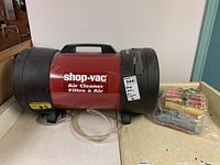 Shop Vac air cleaner unit with power cord, red and black color, on counter with extra filters in plastic bag adjacent.