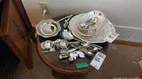 Overview of the collection of silver plated serving pieces and utensils on a round table, showing trays, spoons, and other items.