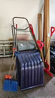 Wide view showing a large snow pusher with a wide blue plastic blade and metal frame, standing alongside several red-handled snow shovels of various designs and sizes in a garage area.