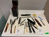 Full view of assorted knives, fillet tools laid out on white counter, including sheaths and wooden storage box