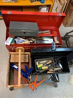 Overall view of the lot showing red metal toolbox, wooden tote with clamps, and black metal toolbox with various small hand tools.