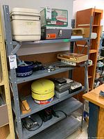 Metal shelving unit with various toolboxes, router bit cases, tape measures, and power tools including Craftsman palm sander.