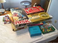 Stack of multiple vintage board games including Risk, Scrabble, Clue, Farming Game Kids, Trivial Pursuit, and Rummoli, showing titles on box tops.