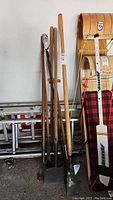 Full view of all yard tools with wooden handles leaning against a wall and ladder in background