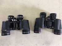 Two black binoculars side by side on a light surface, showing general shape and size.