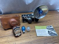 Argus C3 camera with attached flash and reflector, leather case, light meter, manual, and bulbs arranged on a wooden surface.