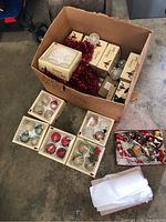 Box with multiple boxed and loose Christmas ornaments including red tinsel garland, and small tray with holiday items.
