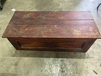 Top down view of the wooden trunk showing worn reddish-brown wood, uneven top planks, and carved handle on front panel.