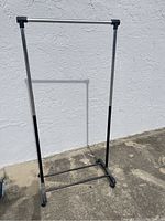 Full view of lightweight metal clothing rack with black plastic connectors and adjustable height poles against a white wall outdoors.