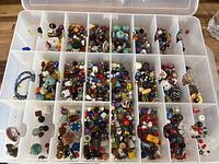 Top view of open clear plastic storage box with multiple compartments containing a large assortment of colorful crafting beads in various shapes and sizes, including some flower-shaped and patterned beads.