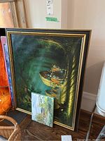 Framed large dark-toned painting on canvas with figures and natural scenery leaning against wall, with smaller paintings stacked in front