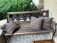 Front-facing view of the outdoor bench showing the wood slatted seat and decorative metal leaf backrest with three striped cushions placed on and beside the bench.