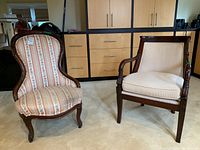 Both chairs shown side by side in a room with wood cabinetry in background. The armchair on the right has cream upholstery and carved armrests and legs. The balloon back chair on the left has floral striped upholstery and curved legs.