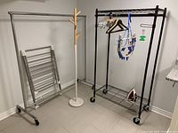 Overview photo showing all items in the lot grouped together in a corner: two metal racks (one chrome, one black), wooden coat rack, drying rack against the wall, T-fal iron on bottom shelf, various hangers hanging or resting.