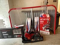 Photo showing Sherwood hockey net standing with white netting, red frame. CCM goalie pads, gloves, helmet with cage, and hockey sticks in front.