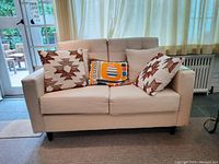 Front view of beige upholstered love seat with five throw pillows showing geometric and solid patterns.