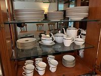 Glass-front wooden china cabinet shelf holding stacks of plates including dinner plates, side plates, bowls, teapot, sugar bowl, creamer, cups and saucers