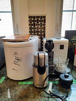 Three appliances on kitchen counter: white Charles Craft bread maker, white and black Nespresso DeLonghi coffee maker with two glass espresso cups on saucers, KitchenAid stainless steel electric coffee grinder in front