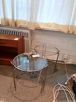 Two round metal framed glass top tables positioned near a window and on a tiled floor, showing their size and general condition.